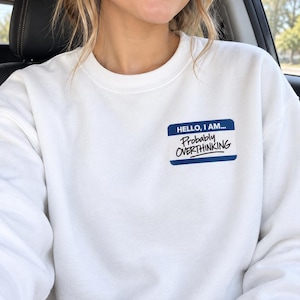 Puede incluir: Una sudadera blanca de cuello redondo con un gráfico rectangular de etiqueta de nombre en azul y blanco en el pecho. El gráfico incluye el texto HELLO, I AM... en letras azules y el texto negro manuscrito Probably OVERTHINKING debajo.