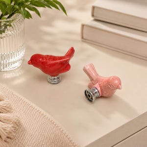 Könnte beinhalten: Zwei vogelförmige Schrankknöpfe, einer in leuchtendem Rot und der andere in zartem Pastellrosa. Jeder Knopf besteht aus einer glänzenden Keramikvogel-Figur, die auf einem runden, silberfarbenen Metallsockel mit Gewindeanschluss montiert ist.