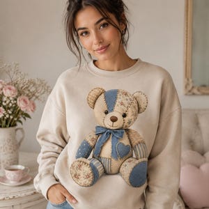 May include: A beige crewneck sweatshirt featuring a large, centred patchwork teddy bear graphic. The bear is constructed from various blue and floral-patterned fabric segments, accented with a blue bow and a small blue heart on its chest.