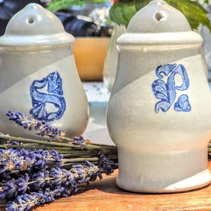 May include: Two off-white ceramic salt and pepper shakers with a smooth, rounded shape. Each shaker features a single blue hand-painted letter, with one displaying an S and the other a P, both in a decorative, stylized font with floral accents.