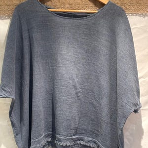 May include: A slate blue, loose-fitting top with a wide, rounded neckline and short, wide sleeves. The garment features a textured, woven fabric appearance and a frayed, raw-edge hemline along the bottom and sleeve cuffs, hanging on a wooden hanger.