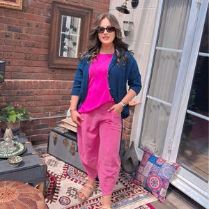 May include: A bright pink top and matching pink trousers paired with a dark blue button-down shirt. The outfit includes beige strappy sandals and black sunglasses. The clothing features a relaxed fit with visible pockets on the trousers.
