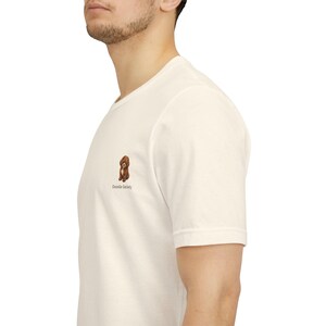 Könnte beinhalten: Ein cremefarbenes Kurzarm-T-Shirt mit einer kleinen gestickten Grafik eines braunen, lockigen Hundes auf der linken Brustseite. Unter der Hundeillustration ist der Text "Doodle Society" in einer einfachen, dunklen Schrift gedruckt.