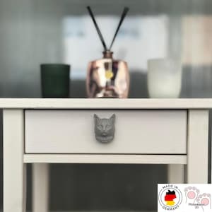 Könnte beinhalten: Ein hellgrauer Möbelknopf in Form eines Katzenkopfes, der an der Vorderseite einer weißen Holzschublade befestigt ist. Der Katzenknopf zeigt detaillierte Gesichtszüge, einschließlich spitzer Ohren, Augen und Schnurrhaare als Griff.