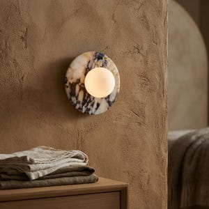 May include: A circular wall lamp featuring a polished marble base with white, grey, and dark blue veining. A spherical, frosted white glass globe is mounted off-center on the marble disc, emitting a soft, warm glow against the textured wall.