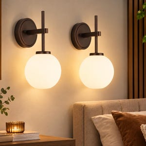 May include: A pair of wall-mounted light fixtures featuring dark bronze-toned metal hardware and circular backplates. Each fixture has a vertical stem supporting a spherical, frosted white glass globe shade that emits a warm, soft glow.