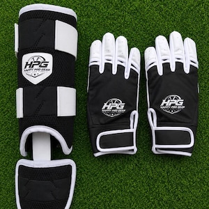 Puede incluir: Una espinillera en blanco y negro y un par de guantes a juego. La espinillera cuenta con correas blancas y un logotipo que dice HPG HAPPY PRO GEAR. Los guantes tienen dedos blancos, palmas negras y el mismo logotipo HPG HAPPY PRO GEAR.