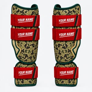 Puede incluir: Un par de espinilleras con un patrón de camuflaje verde y marrón y ribete verde oscuro. Cada espinillera tiene tres correas horizontales rojas con el texto blanco YOUR NAME impreso en cada correa en una fuente sans-serif en negrita.