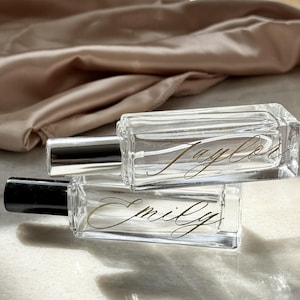 May include: Two rectangular clear glass perfume bottles with black spray caps. The top bottle features the name Taylor in gold cursive script, and the bottom bottle features the name Emily in gold cursive script, both printed on the glass surface.