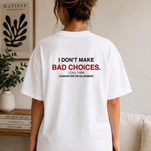 Peut inclure: Un t-shirt blanc à manches courtes avec un texte noir et rouge imprimé au dos : I DON'T MAKE BAD CHOICES. I CALL THEM CHARACTER DEVELOPMENT. Le vêtement présente une encolure ronde et une coupe décontractée, mettant en valeur le message.