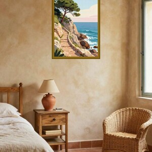 Könnte beinhalten: Ein gerahmter Kunstdruck mit einer Küstenlandschaft, einem steinernen Pfad entlang felsiger Klippen, einer grünen Kiefer und einem blauen Ozean unter einem hellen Himmel. Das Bild hängt an einer strukturierten beigen Wand über einem Nachttisch.
