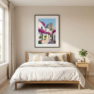 Könnte beinhalten: Ein gerahmter Kunstdruck, der eine mediterrane Straßenszene mit weißen Gebäuden, leuchtend rosa Bougainvillea-Blüten, grünen Topfpflanzen und einem felsigen Berggipfel unter einem klaren blauen Himmel zeigt, präsentiert in einem Schlafzimmer.