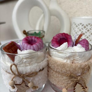 Puede incluir: Dos velas en vaso decoradas con nata montada de cera blanca, frambuesas de cera morada y cuadrados de chocolate de cera marrón. Cada vaso tiene una funda de tela texturizada atada con cuerda y pequeños adornos de madera en forma de estrella y árbol.