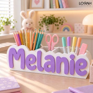 Puede incluir: Un organizador de escritorio blanco con el nombre Melanie en letras grandes de color morado en la parte frontal. El organizador contiene una colección de lápices de colores, unas tijeras rosas, varios rotuladores azules, morados y amarillos, y una goma rosa.