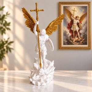 May include: A white and gold statue of an archangel standing on a rocky base, holding a tall gold cross-topped staff and a shield. The figure features large, detailed gold wings and is depicted in a dynamic pose, with a framed print behind it.