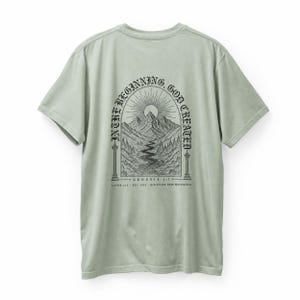 May include: A sage green short-sleeved t-shirt featuring a black ink-style graphic on the back. The design depicts a mountain landscape under a sunburst, framed by an archway with the text "IN THE BEGINNING, GOD CREATED" and "GENESIS 1:1".