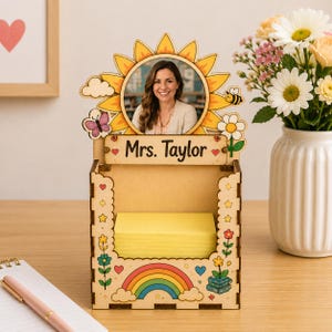Könnte beinhalten: Ein Schreibtisch-Organizer aus Holz mit einem sonnenblumenförmigen Rahmen, einem runden Foto einer Person, dem Schriftzug "Mrs. Taylor" auf einem Schild und einem Stapel gelber Haftnotizen. Die Basis ist mit bunten Blumen, einem Regenbogen und einer Biene verziert.