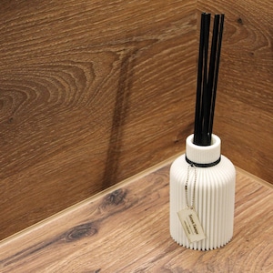 May include: A white cylindrical reed diffuser with a vertical ribbed texture and a narrow neck. It contains several thin black reeds extending upwards. A small rectangular tag with the text "Sweet Tange" hangs from a silver ball chain around the neck.