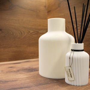 May include: A large white ribbed vase and a smaller white ribbed reed diffuser bottle with black reeds. The smaller bottle features a black neck band and a rectangular tag with the text Secret Escape attached by a silver ball chain.