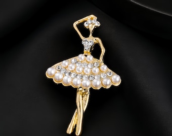 May include: A gold-toned metal brooch shaped like a ballerina. The figure features a bodice and tutu skirt embellished with clear round rhinestones and white faux pearls. The ballerina is posed with one arm raised above the head and legs crossed.