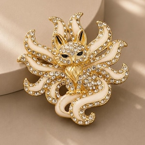 Può includere: Una spilla color oro a forma di volpe a nove code, caratterizzata da inserti in smalto bianco e strass trasparenti scintillanti incastonati su tutto il corpo e le code. La volpe ha occhi neri e un petto dorato testurizzato, con finitura lucida.