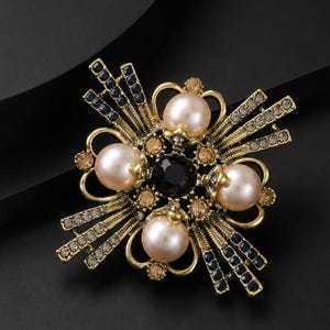 May include: A gold-tone brooch featuring four large, round, cream-colored faux pearls arranged in a cross pattern around a central black faceted stone. The design includes radiating metal rays accented with small dark blue and champagne-colored crystals.