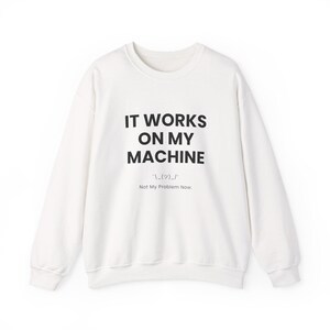 May include: A white long-sleeve sweatshirt featuring black text that reads IT WORKS ON MY MACHINE, followed by a shrug emoticon and the smaller text Not My Problem Now. The garment has a crew neckline and ribbed cuffs at the sleeves and hem.