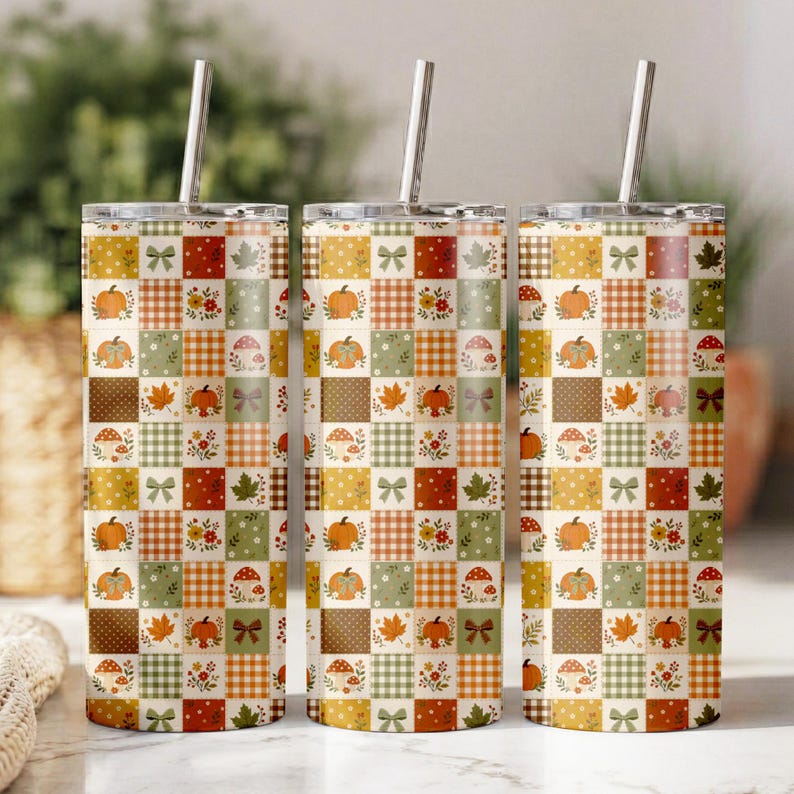 May include: Three cylindrical tumblers with metal straws, each featuring a patchwork pattern of squares in shades of orange, green, brown, and white. The design includes motifs of pumpkins, mushrooms, autumn leaves, flowers, and bows.