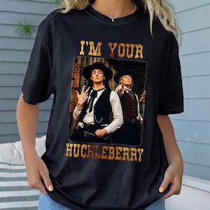 May include: A black short-sleeve t-shirt featuring a graphic print of two individuals in western attire with vests and hats. The text "I'M YOUR HUCKLEBERRY" is printed in a distressed gold font above and below the central photographic image.