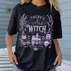 May include: A black short-sleeve t-shirt featuring a light purple graphic print. The design includes the text "THERE'S A LITTLE WITCH" and "THE OWENS SISTERS" alongside illustrations of a house, a lavender bottle, a tequila bottle, and a bowl.