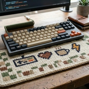 May include: A rectangular, cream-coloured crocheted keyboard wrist rest featuring pixelated motifs including a game controller, a heart, a coffee mug, and a lightning bolt, with a checkered border in shades of green and brown.