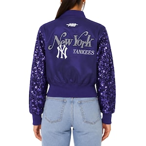 Puede incluir: Una chaqueta bomber morada con mangas largas cubiertas de lentejuelas moradas a juego. La parte trasera presenta el texto New York en purpurina plateada, el logotipo blanco entrelazado de los NY Yankees y un pequeño logotipo blanco Pro en el centro del cuello.