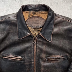 May include: A dark brown distressed leather jacket with a pointed collar and a central metal zipper. The interior lining features a tan fabric with a printed pattern and a leather patch label that reads Original Leather Wear TRAPPER.