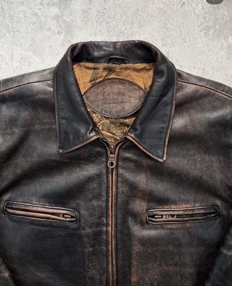 May include: A dark brown distressed leather jacket with a pointed collar and a central metal zipper. The interior lining features a tan fabric with a printed pattern and a leather patch label that reads Original Leather Wear TRAPPER.