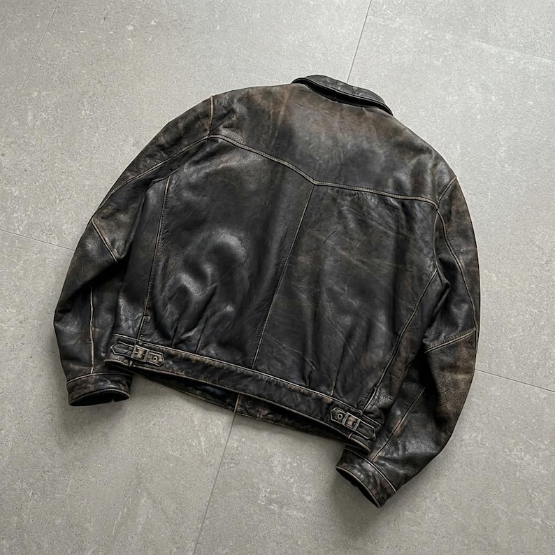 May include: A dark brown leather jacket featuring a distressed finish, a classic collar, long sleeves, and a back yoke. The hem includes adjustable side straps with metal buckles, providing a structured fit for this durable outer garment.