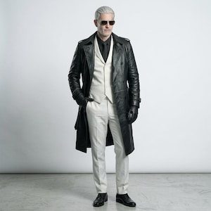 Pode incluir: Um sobretudo de couro preto usado sobre um colete branco, calças brancas e uma camisa preta. O traje inclui luvas de couro preto, sapatos sociais pretos e óculos de sol escuros. O casaco apresenta um design de peito duplo com lapelas.