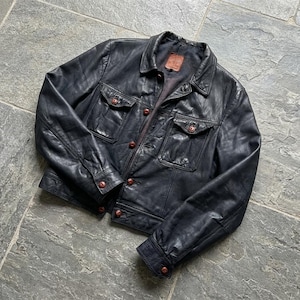 May include: A dark navy blue leather jacket featuring a classic collar, long sleeves with buttoned cuffs, and two chest pockets with button-down flaps. The front closure consists of a vertical row of brown buttons, and a brown label is at the neck.