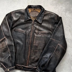 May include: A dark brown leather jacket featuring a distressed finish, a central zipper closure, a pointed collar, and two horizontal zippered chest pockets. The cuffs and hem include snap button closures, and the interior lining is tan.