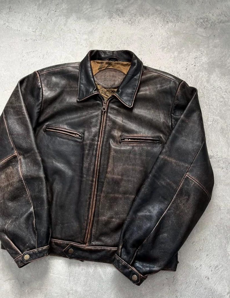 May include: A dark brown leather jacket featuring a distressed finish, a central zipper closure, a pointed collar, and two horizontal zippered chest pockets. The cuffs and hem include snap button closures, and the interior lining is tan.