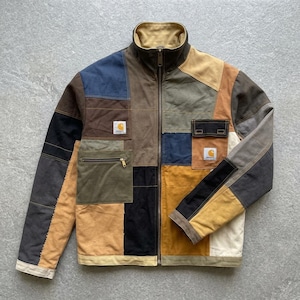 May include: A full-zip jacket constructed from a patchwork of multi-coloured canvas panels in shades of brown, tan, navy, black, and olive green. The jacket features a stand-up collar, a zippered chest pocket, a flap pocket, and two Carhartt labels.