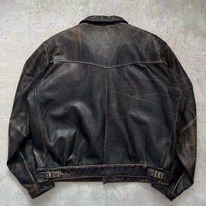 May include: A dark brown leather jacket featuring a pointed collar, a horizontal back yoke seam, and adjustable side buckle straps at the hem. The leather displays a distressed, worn-in finish with visible variations in color and texture throughout.
