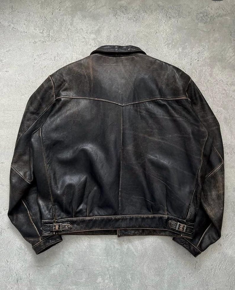 May include: A dark brown leather jacket featuring a pointed collar, a horizontal back yoke seam, and adjustable side buckle straps at the hem. The leather displays a distressed, worn-in finish with visible variations in color and texture throughout.