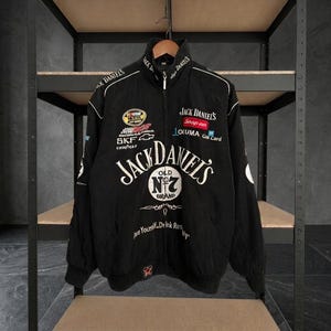 May include: A black racing-style jacket featuring a high collar and full-length front zipper. The front displays multiple embroidered logos including Jack Daniel's, Snap-on, and Chevrolet, with the text "Pace Yourself. Drink Responsibly" at the base.