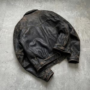 May include: A dark brown distressed leather jacket featuring a folded collar, long sleeves with buttoned cuffs, and adjustable side straps with metal buckles at the hem. The jacket displays a weathered texture with visible seams and a back yoke.