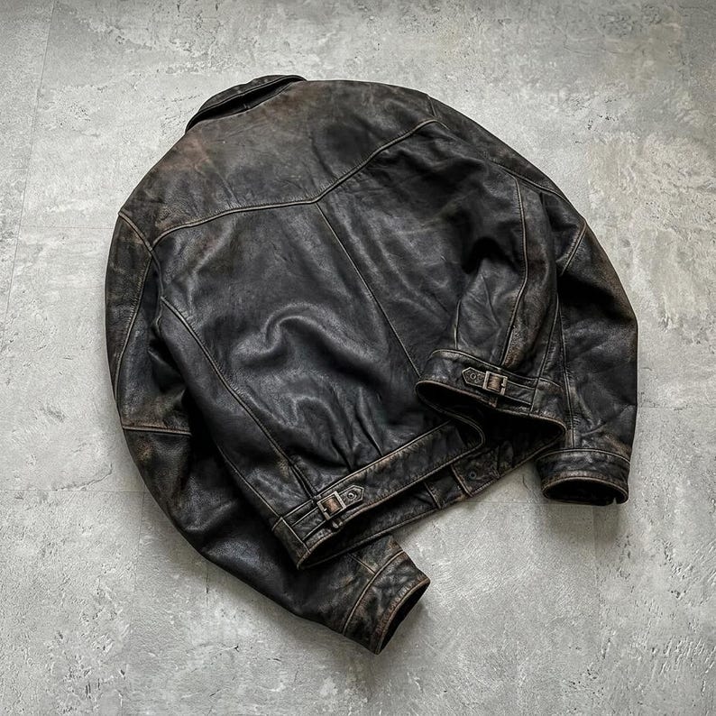 May include: A dark brown distressed leather jacket featuring a folded collar, long sleeves with buttoned cuffs, and adjustable side straps with metal buckles at the hem. The jacket displays a weathered texture with visible seams and a back yoke.