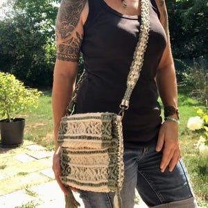 May include: A rectangular macrame crossbody bag featuring a geometric pattern in sage green and off-white cord. The bag includes a matching braided shoulder strap, a fold-over flap, and decorative fringe tassels hanging from the bottom corners.