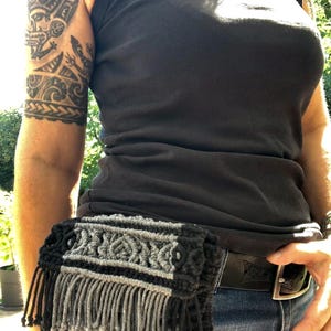 May include: A rectangular macrame waist bag featuring a repeating geometric knot pattern in alternating black and grey cord. The bag includes a decorative fringe detail along the centre section and is attached to a black leather belt with a metal buckle.