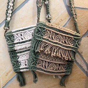 May include: Two macrame crossbody bags featuring intricate knotted patterns and fringe details. One bag combines sage green and cream cord, while the other features sage green and tan cord. Both bags include matching macrame shoulder straps.