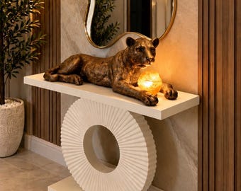 Puede incluir: Una mesa consola blanca con base circular estriada sostiene una escultura de pantera color bronce y una esfera ámbar brillante. Sobre la mesa cuelga un espejo irregular con borde dorado y un reloj de pared con números romanos negros.