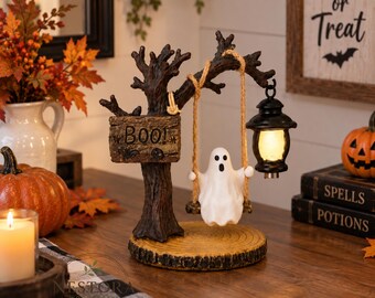 Puede incluir: Un adorno decorativo de Halloween para mesa que presenta una figura de fantasma blanco sentada en un columpio suspendido de una rama de árbol marrón oscuro. El árbol tiene un letrero que dice BOO! y una linterna negra. La base es de madera.